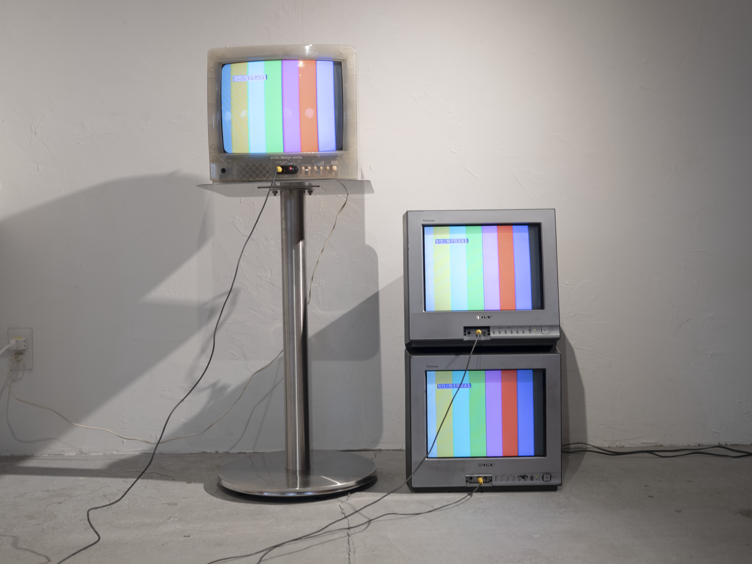 一本足テーブルの上と床に、3台のブラウン管テレビを配置した写真。白いブラウン管テレビ1台と、グレーのビデオモニター2台が並び、3台ともカラーバーを映している。
